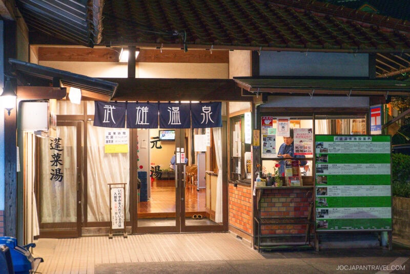 A traditional Japanese onsen entrance at night, warmly lit with wooden pillars, banners with Japanese characters, and an information board—evoking the timeless charm of Takeo Onsen.