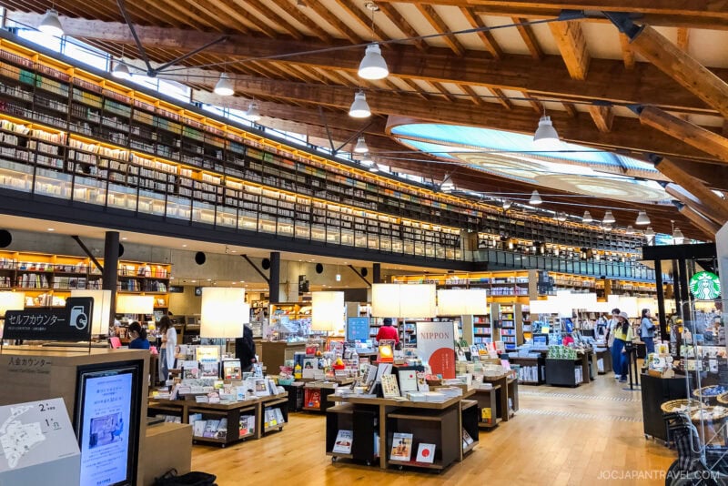 A spacious, modern bookstore inspired by Takeo Onsen, with wooden beams, high ceilings, and large windows. Bookshelves line the upper walls while tables display books and magazines as people browse under warm lighting.