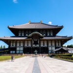 Todaiji Temple (東大寺) – Nara’s Great Buddha and Historic Temple The image shows Todaiji Temple in Nara, Japan—a large, historic wooden building with a wide tiled roof—set against a clear blue sky, with a few visitors walking in the foreground.