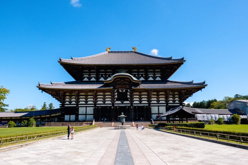 The image shows Todaiji Temple in Nara, Japan—a large, historic wooden building with a wide tiled roof—set against a clear blue sky, with a few visitors walking in the foreground.