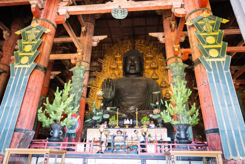 Large bronze statue of Buddha seated with one hand raised at Todaiji, surrounded by an elaborate golden backdrop and ornate wooden columns, with greenery and offerings placed in the foreground.