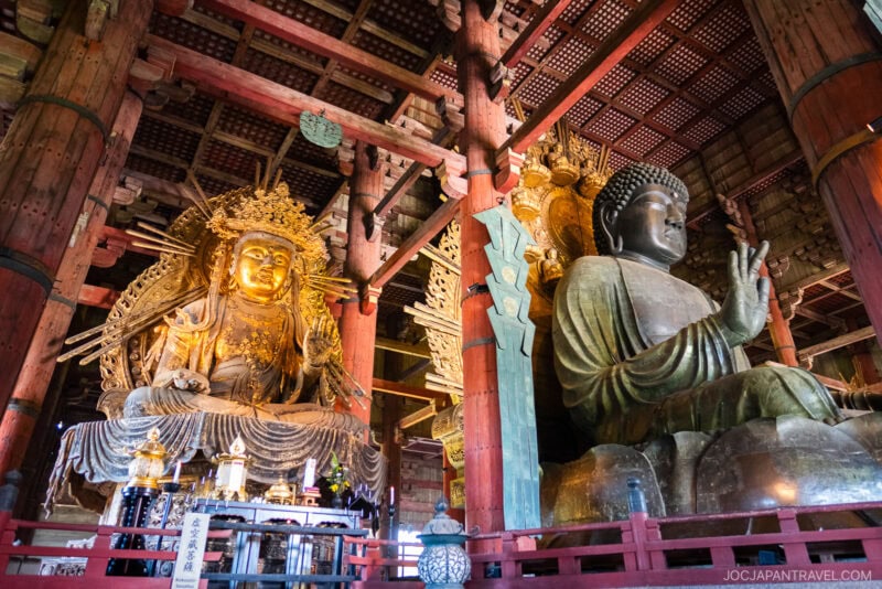The large bronze Buddha statue and ornate golden statue inside Todaiji Temple in Nara, Japan, are surrounded by wooden pillars and intricate architectural details.