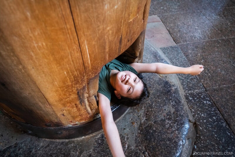 A smiling child squeezes through a large wooden pillar at Todaiji, arms outstretched while lying on a stone floor in a brightly lit indoor setting.