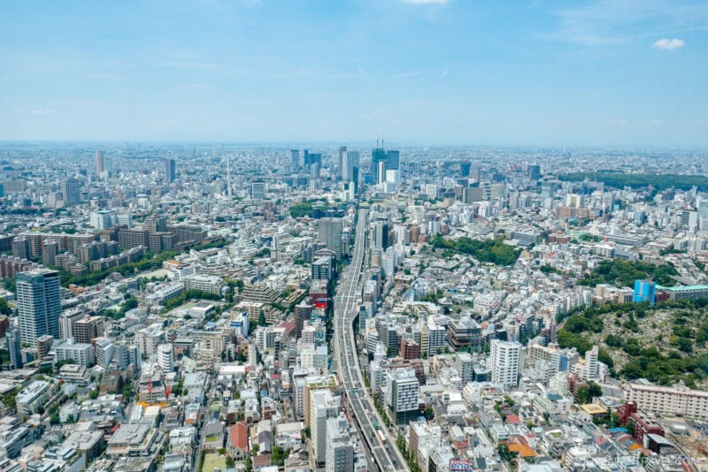 Aerial view of a sprawling Tokyo city view with numerous buildings, roads, and green spaces under a clear blue sky, showcasing a vibrant and densely populated urban environment.