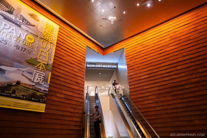 Two people ride escalators in a brightly lit modern building with wooden panel walls. A large poster about Japanese architecture is on the left, and a sign above reads MORI ART MUSEUM, offering a glimpse into the Tokyo City View experience.