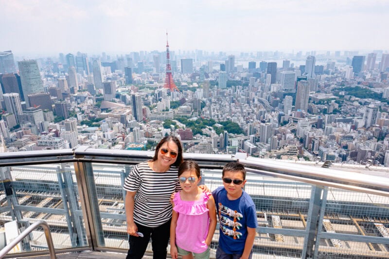 A woman and two children stand on an observation deck, taking in the breathtaking Tokyo city view with Tokyo Tower rising above the sprawling skyline on a bright day.