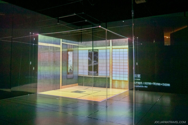A digital art installation displays a traditional Japanese tatami room projected within a transparent cube frame, set against a dark gallery space. The realistic room features shoji screens, art, soft lighting, and hints of a distant Tokyo city view.