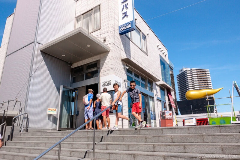 People walk down steps outside the white Tokyo Cruise building on a sunny day; two boys in sunglasses are in the foreground, while others stand near the entrance. A yellow slide and tall buildings add to the lively Tokyo Cruise atmosphere.