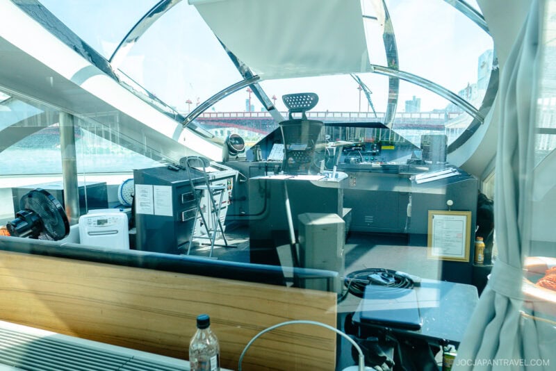 The image shows the control room of a modern Tokyo Cruise boat or ferry, viewed through a window, featuring control panels, equipment, documents, and a cityscape in the background through large windows.