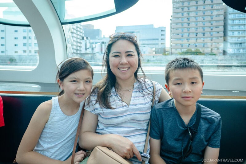 A woman sits between two children, all smiling at the camera, inside a bright, modern Tokyo cruise boat with city buildings visible through large windows behind them.