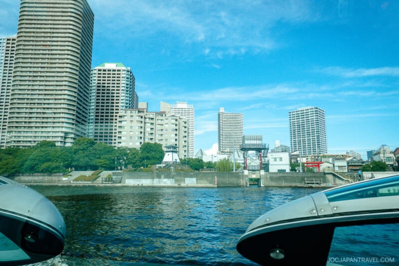 A cityscape featuring tall modern buildings and apartments along a waterfront under a clear blue sky, with water and parts of futuristic boats in the foreground, capturing the vibrant energy of a Tokyo cruise.