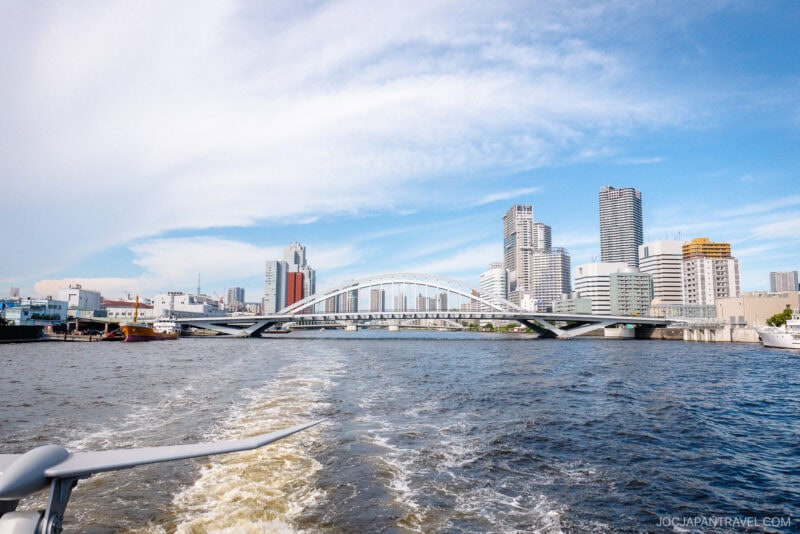 City skyline with modern high-rise buildings and a white arch bridge spanning a wide river, viewed from a Tokyo cruise boat leaving a foamy wake under a blue sky with scattered clouds.