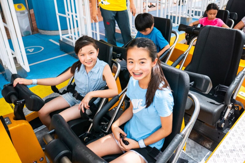 Two smiling girls sit side by side in a roller coaster car at Tokyo Dome City, ready for the ride. Other children are seated behind them, and a person in a yellow shirt stands nearby. The setting is bright and cheerful.