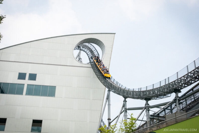 A yellow roller coaster car filled with people descends a steep track through a circular opening in a modern white building at Tokyo Dome City. The sky is clear, and some greenery is visible below.