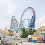 Tokyo Dome City (東京ドームシティ) – Family Fun in the Heart of Tokyo A bright, sunny day at an amusement park like Tokyo Dome City with large, looping roller coasters and a Ferris wheel. Colorful signs, information booths, and a restroom area are visible in the foreground.
