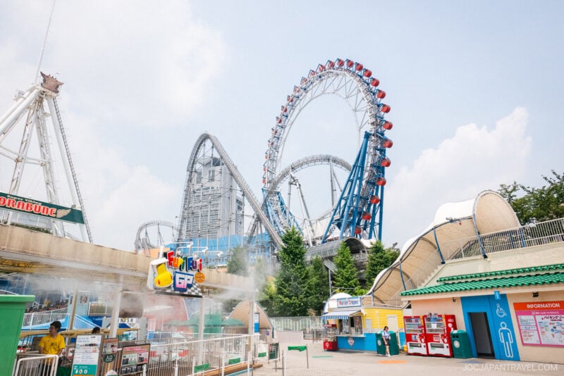 A bright, sunny day at an amusement park like Tokyo Dome City with large, looping roller coasters and a Ferris wheel. Colorful signs, information booths, and a restroom area are visible in the foreground.
