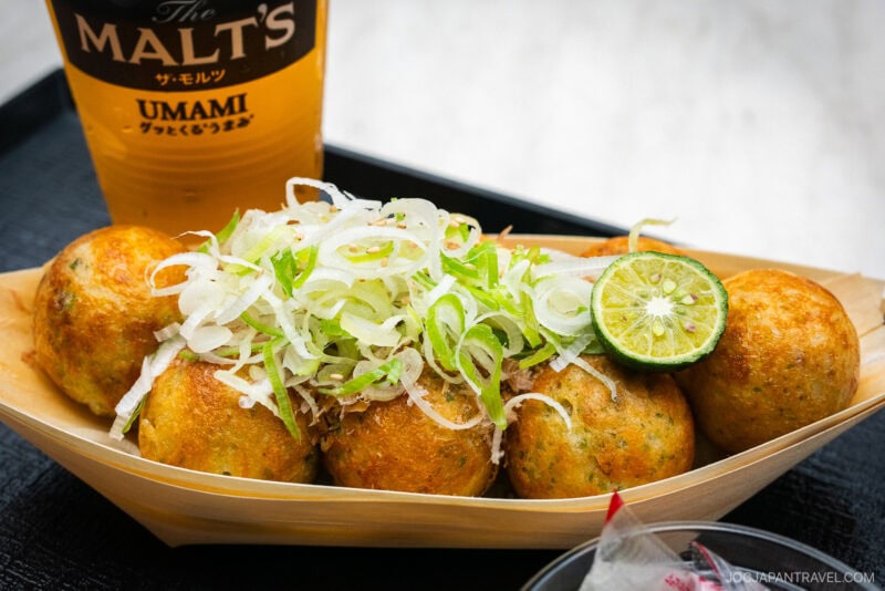 A tray of golden-brown takoyaki balls topped with sliced green onions and a halved lime, served next to a glass of THE MALTS UMAMI beer—perfect for savoring after an exciting day at Tokyo Dome City.