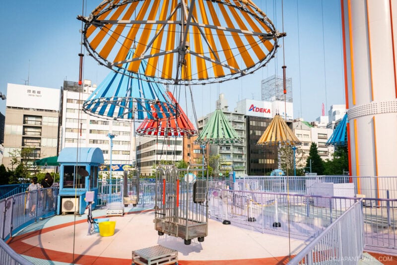 A colorful amusement park ride with striped umbrellas and empty seats hangs above a circular platform, surrounded by blue railings at Tokyo Dome City. City buildings and signs are visible in the background under a clear sky.