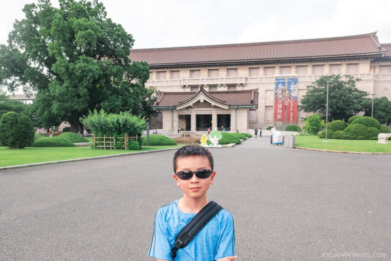 A young boy wearing sunglasses and a blue shirt stands in front of the Tokyo National Museum, with its historic building, green trees, and manicured lawns creating a scenic background.