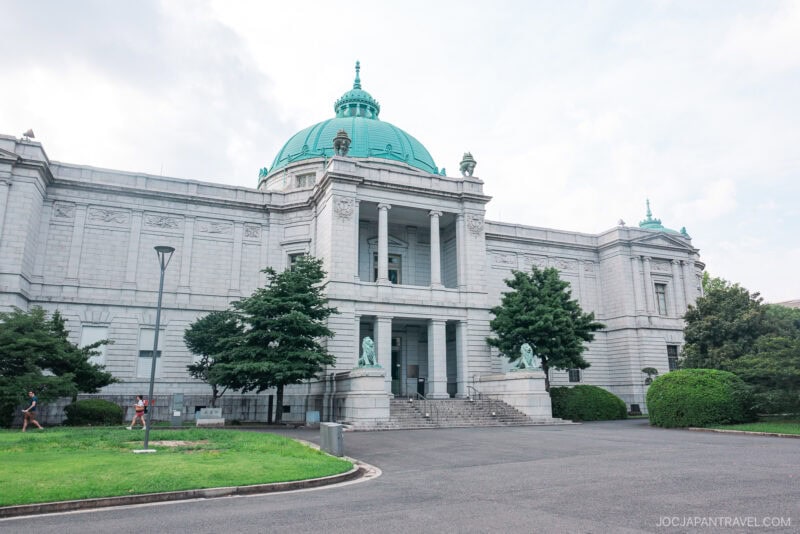 A grand neoclassical building with a green dome, columns, and statues at the entrance stands amid trees and shrubbery under a partly cloudy sky, reminiscent of the Tokyo National Museum’s iconic architecture.