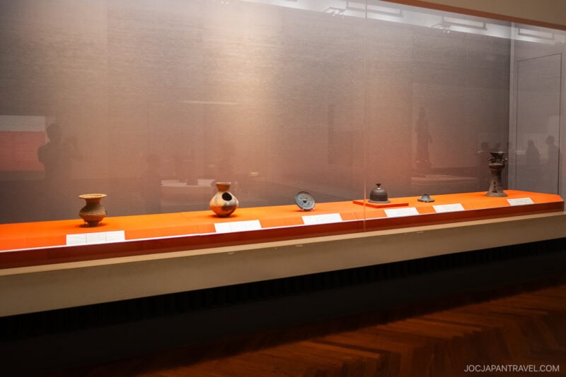 A museum display case at the Tokyo National Museum showcases six ancient pottery artifacts on an orange surface, each labeled. The dimly lit background and glass reflections enhance the exhibit's intriguing atmosphere.