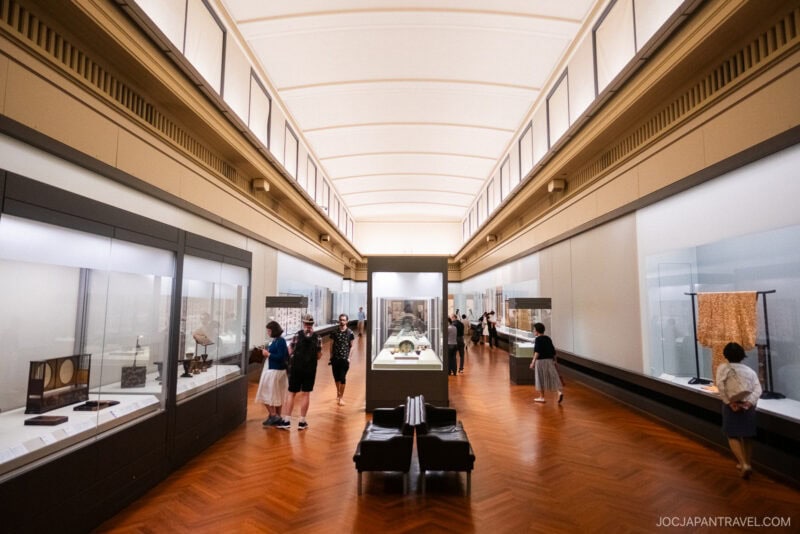 People view artifacts and textiles displayed in glass cases along the walls of a spacious, well-lit Tokyo National Museum gallery with a high ceiling and wooden floor.