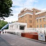 Tokyo National Museum of Nature and Science (国立科学博物館) People walk near the main entrance of the Tokyo National Museum of Nature and Science, a large historic brick building with white stone features. A life-sized blue whale statue stands on the left, while trees border the walkway.