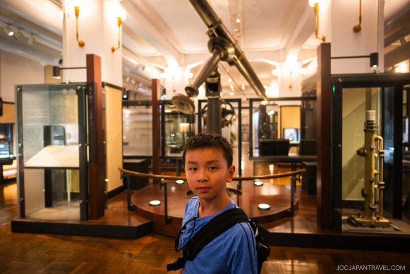 A young boy wearing a blue shirt and backpack stands in the Tokyo National Museum of Nature and Science, surrounded by scientific displays and a large telescope in the background, illuminated by warm lighting.