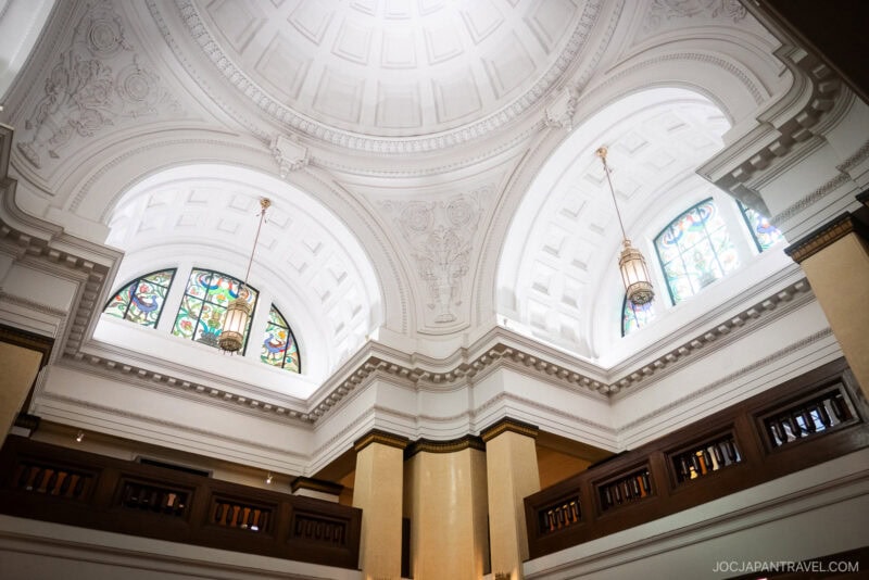 A grand, white-domed ceiling with intricate molding, arched stained glass windows, and hanging lanterns above a balcony with wooden railings in the elegant, historic Tokyo National Museum of Nature and Science.