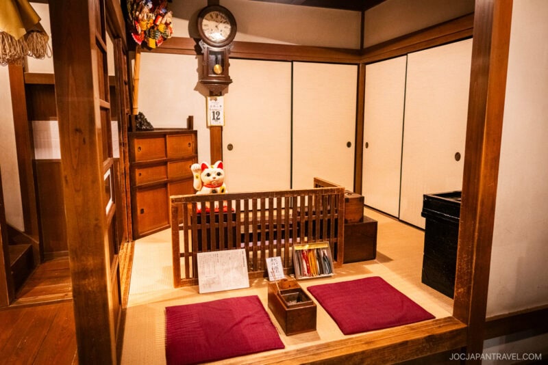 A traditional Japanese room in the Tokyo Shitamachi Museum features tatami floors, sliding shoji doors, a wooden clock, a Maneki-neko figurine, low cushions, and various wooden boxes and shelves.
