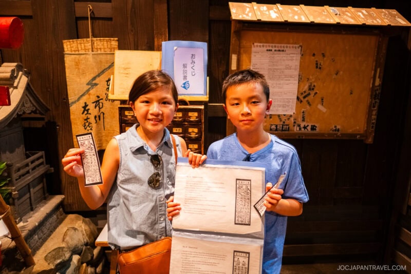 Two children stand indoors at the Tokyo Shitamachi Museum, smiling and holding up papers with printed text and images. Behind them are wooden boards with Japanese writing and a small shrine-like structure, creating a cultural or educational atmosphere.