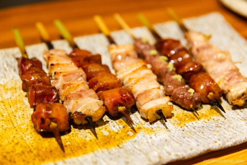 Several skewers of assorted grilled yakitori, including pieces of chicken and vegetables, are arranged on a rectangular ceramic plate, ready to take you down memory lane with every savory bite.