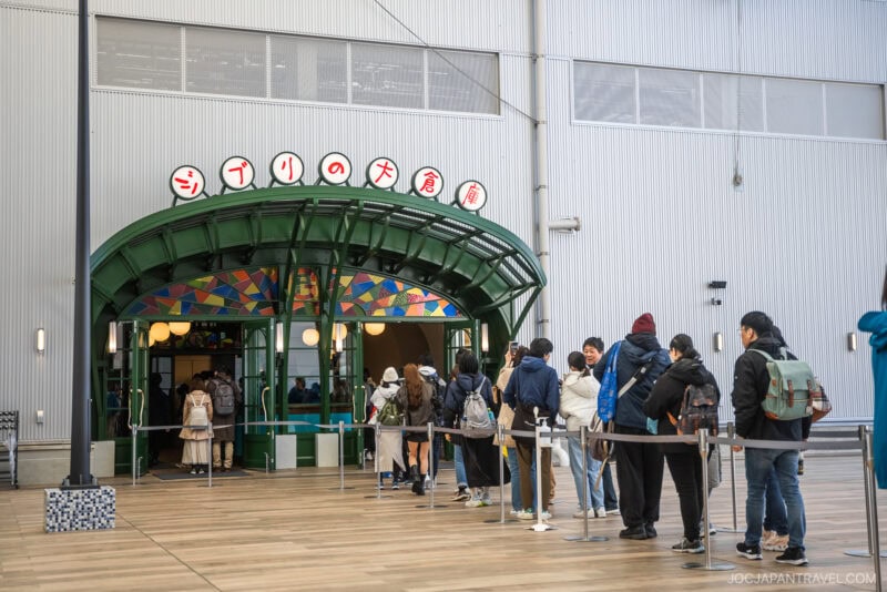 Visitors line up outside a green, arched entrance at Ghibli Park, with colorful Japanese signage above the doorway and a modern wooden deck surrounding the vibrant area.