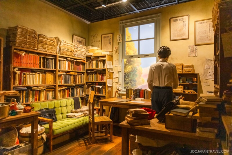 A cozy, cluttered study with wooden shelves filled with books and papers, a green sofa, wooden desks, and a figure by the window as sunlight streams in—evoking the warm, nostalgic atmosphere of Ghibli Park.