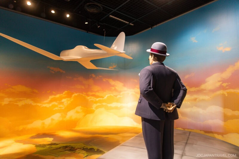 A mannequin in a suit and hat stands with hands behind back, facing a model airplane suspended in a room with a painted sky and clouds, evoking the whimsical atmosphere of flight and imagination found in Ghibli Park.