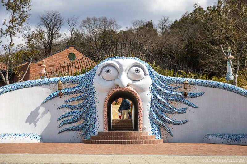 A whimsical sculpture of a face with wide eyes and blue mosaic hair forms an arched tunnel entrance at Ghibli Park, with two people walking through, set against a backdrop of trees and buildings.