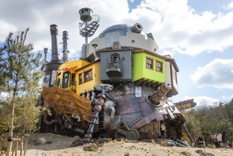 A whimsical, steampunk-inspired building with mechanical legs, gears, and pipes stands like a fantastical moving house—reminiscent of ghibli park—surrounded by trees and people admiring its colorful rooms and balconies.