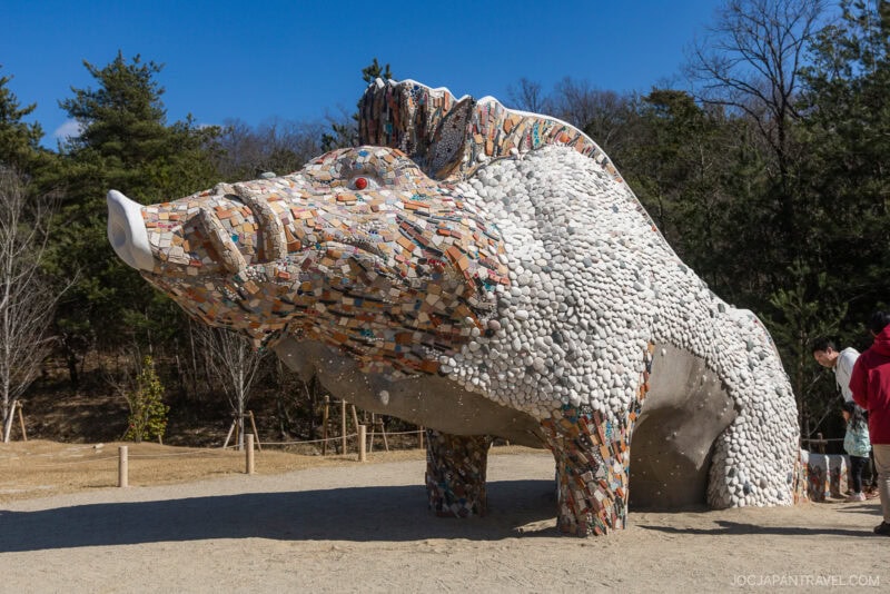 A large boar sculpture covered in colorful mosaic tiles stands outdoors at Ghibli Park on a sunny day, with trees and several people nearby.