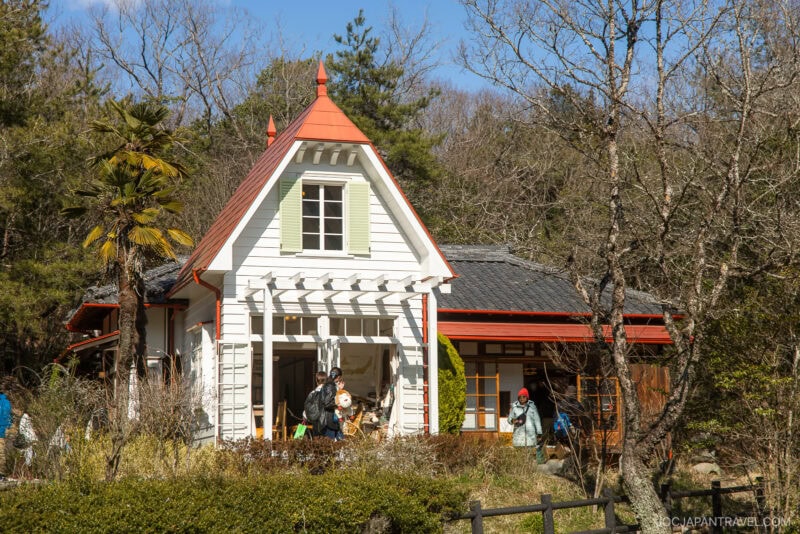 A charming white house with a red roof and green shutters, surrounded by trees and bushes, evokes the whimsical feel of Ghibli Park. People gather at the entrance, enjoying a sunny day at this storybook-inspired building.