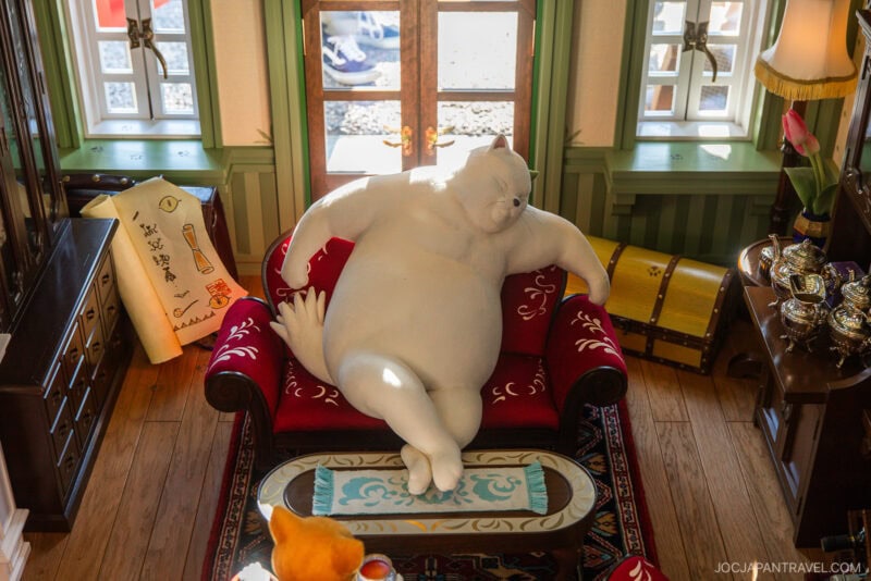 A large, white, relaxed cat figurine lounges on a red velvet sofa in a cozy, sunlit room with wooden floors and green walls—reminiscent of the whimsical charm found in Ghibli Park—adorned with small, detailed decorations and viewed from above.