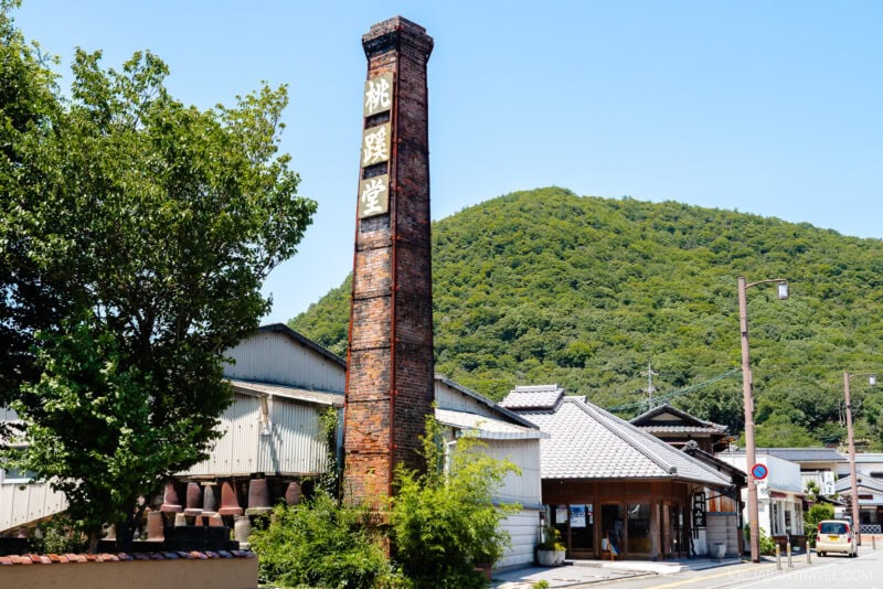 A tall brick chimney with Japanese characters stands beside traditional Japanese buildings, hinting at the legacy of Bizen ware, with a green tree and lush, forested hill in the background under a clear blue sky.
