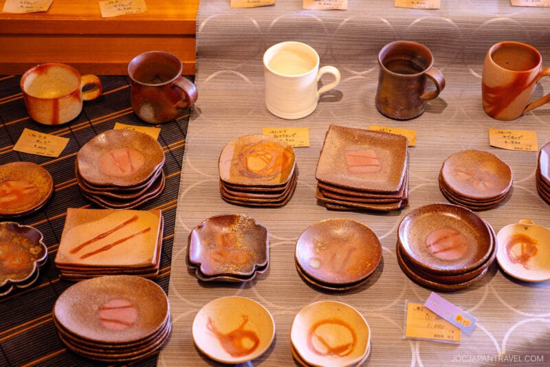 Various handmade ceramic mugs and plates, inspired by bizen ware, are displayed on a table, each with unique earthy tones and patterns. Small handwritten price tags are placed next to the items.