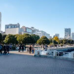 People stroll along the Yamashita Park waterfront promenade, lined with trees and city buildings under a clear blue sky. Some gather in groups, enjoying the scenic views near the water.