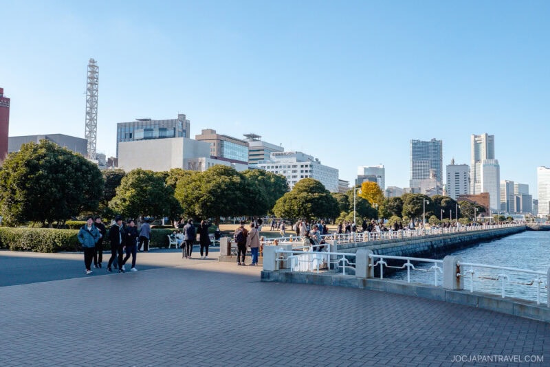 People stroll along the Yamashita Park waterfront promenade, lined with trees and city buildings under a clear blue sky. Some gather in groups, enjoying the scenic views near the water.