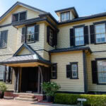 Exploring the Yamate District (山手町) – Yokohama’s Historic Bluff A two-story yellow house with dark shutters and trim, located in the Yamate District, features a covered entryway with columns, several windows, and potted plants by the entrance. The house is surrounded by greenery under a clear blue sky.