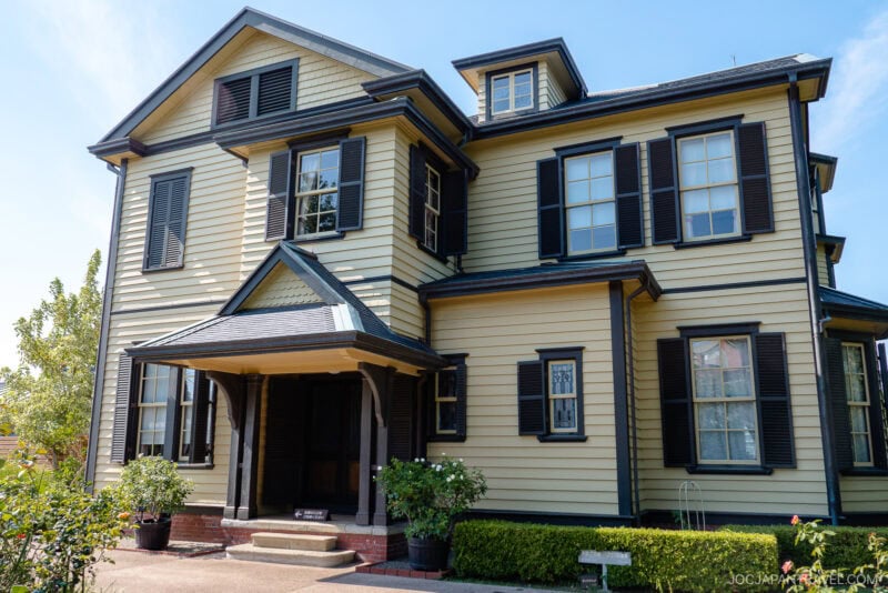 A two-story yellow house with dark shutters and trim, located in the Yamate District, features a covered entryway with columns, several windows, and potted plants by the entrance. The house is surrounded by greenery under a clear blue sky.