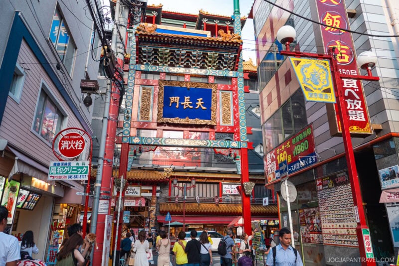 A colorful, ornate Chinese gate marks the entrance to Yokohama Chinatown, where bustling streets lined with shops and bright advertisements invite crowds of people beneath its impressive archway.