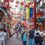 Yokohama Chinatown (横浜中華街) – Japan’s Largest Chinatown A lively street in Yokohama Chinatown is decorated with red lanterns and crowded with people browsing shops and restaurant menus. Colorful signs with Chinese characters line the busy, vibrant scene.