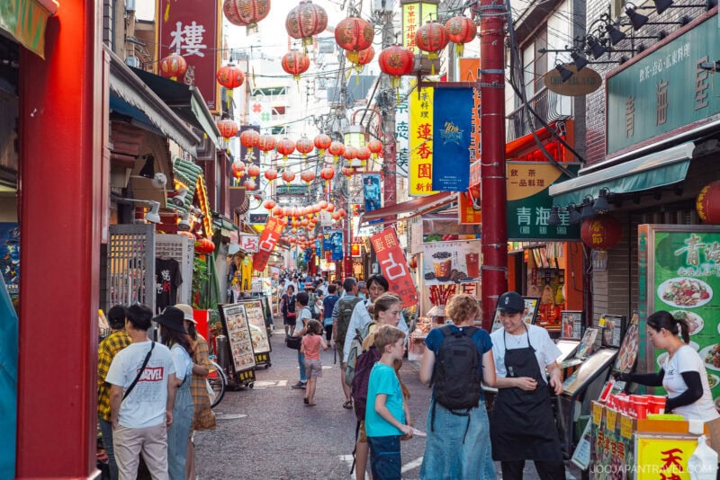 A lively street in Yokohama Chinatown is decorated with red lanterns and crowded with people browsing shops and restaurant menus. Colorful signs with Chinese characters line the busy, vibrant scene.