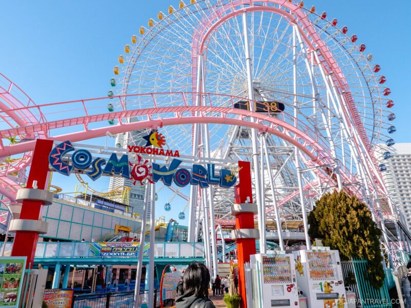 Entrance to Yokohama Cosmo World amusement park with colorful signage welcomes visitors, while the iconic pink Ferris wheel and a thrilling roller coaster stand tall in the background under a clear blue sky.